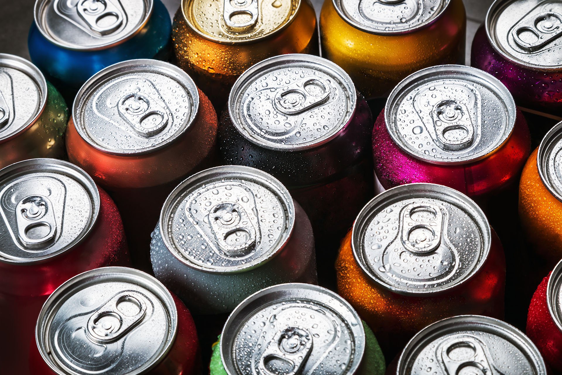 Närbild av olika läskburkar av aluminium med dragflikar, tätt packade tillsammans i en vy uppifrån och ner. Burkarna är i olika starka färger, bland annat rött, orange, blått, grönt, lila och guld, och alla är täckta av kondensdroppar, vilket tyder på att de är kalla och nykylda.