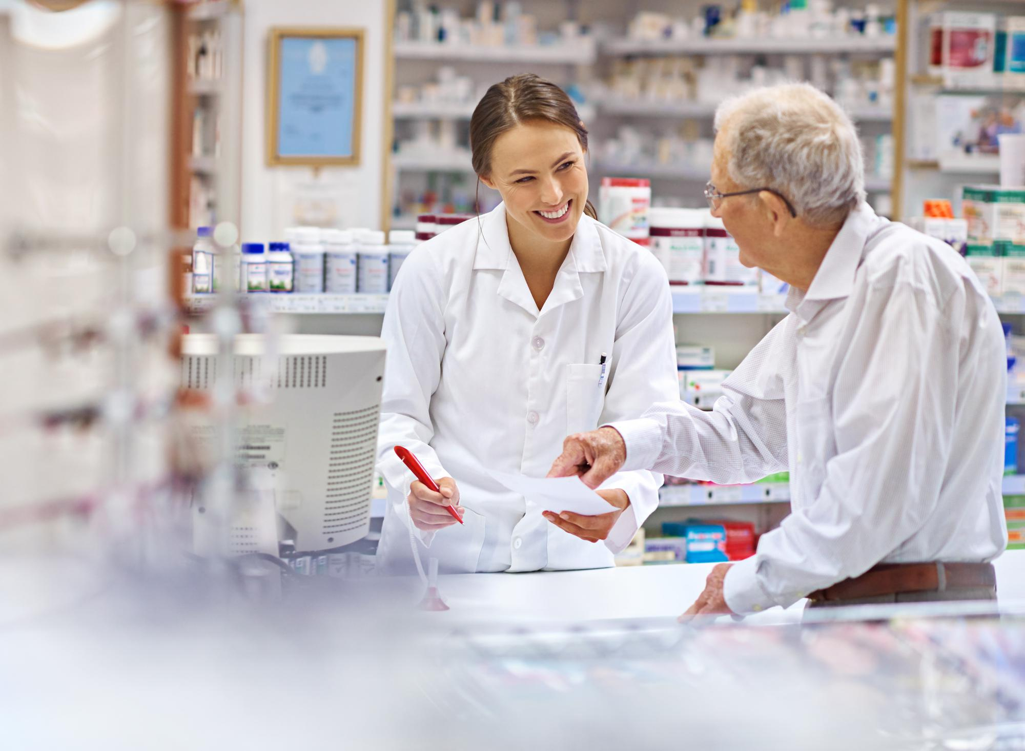 Kvinnlig farmaceut hjälper en äldre manlig kund med ett recept vid en apoteksdisk.