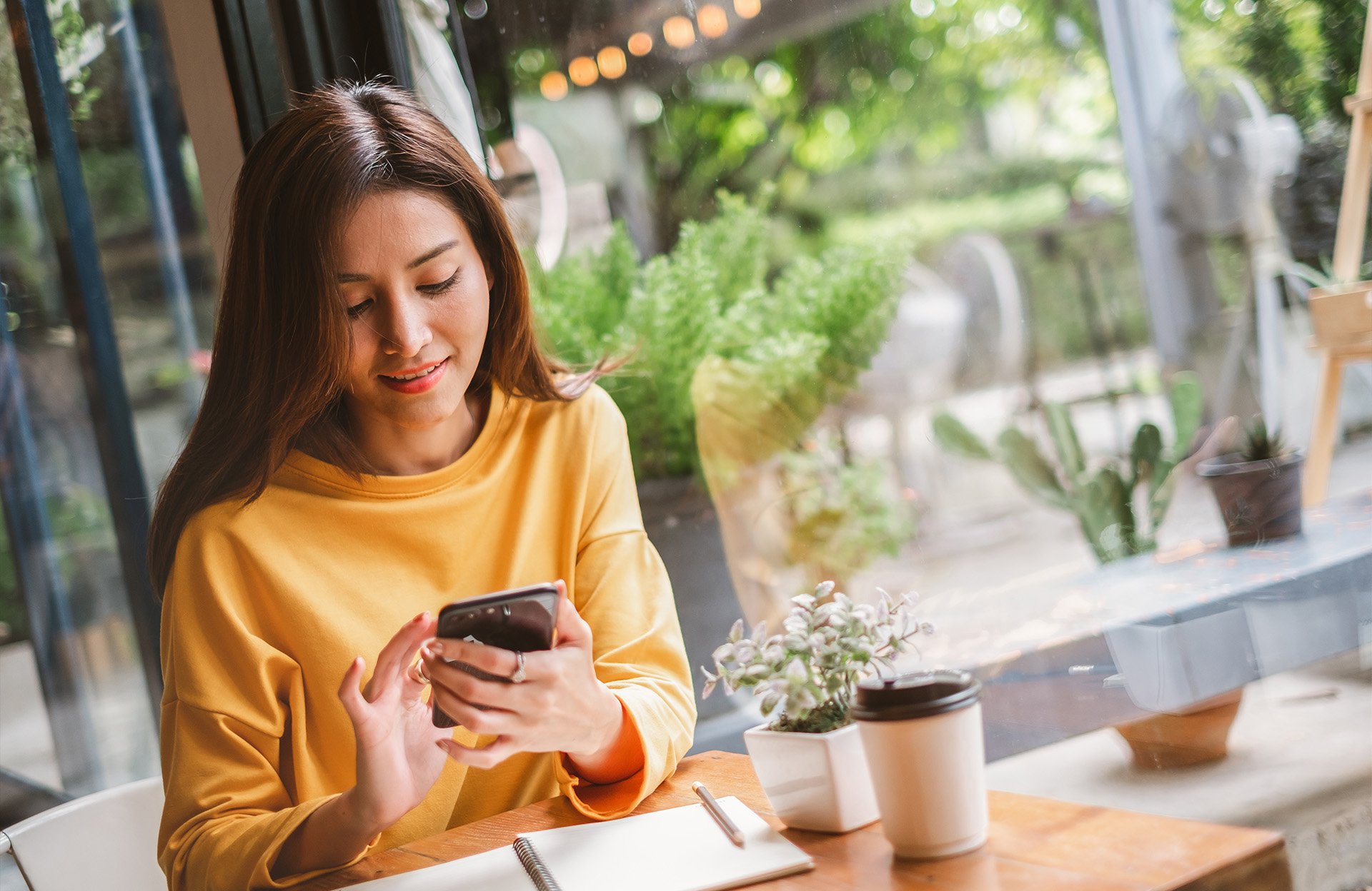 Kvinna som använder en smartphone på ett café med en anteckningsbok och kaffekopp på bordet.