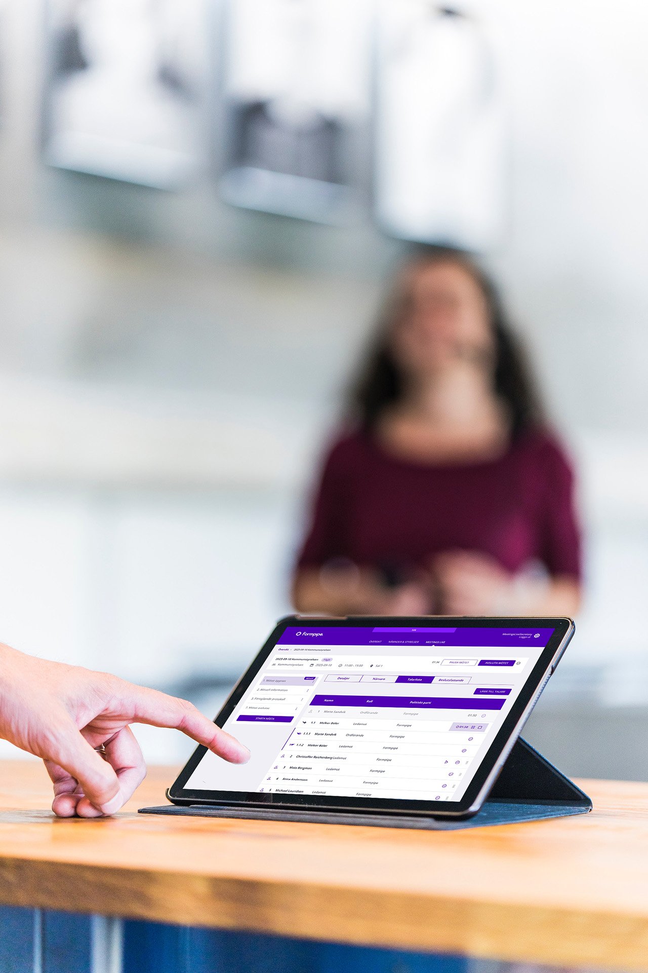 En person pekar på en surfplatta som visar ett detaljerat gränssnitt i Meetings Live från Formpipe, med en suddig gestalt stående i bakgrunden.