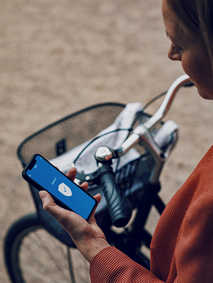 A person holding a smartphone with a blue screen displaying a shield icon, beside a bicycle with a woven basket.