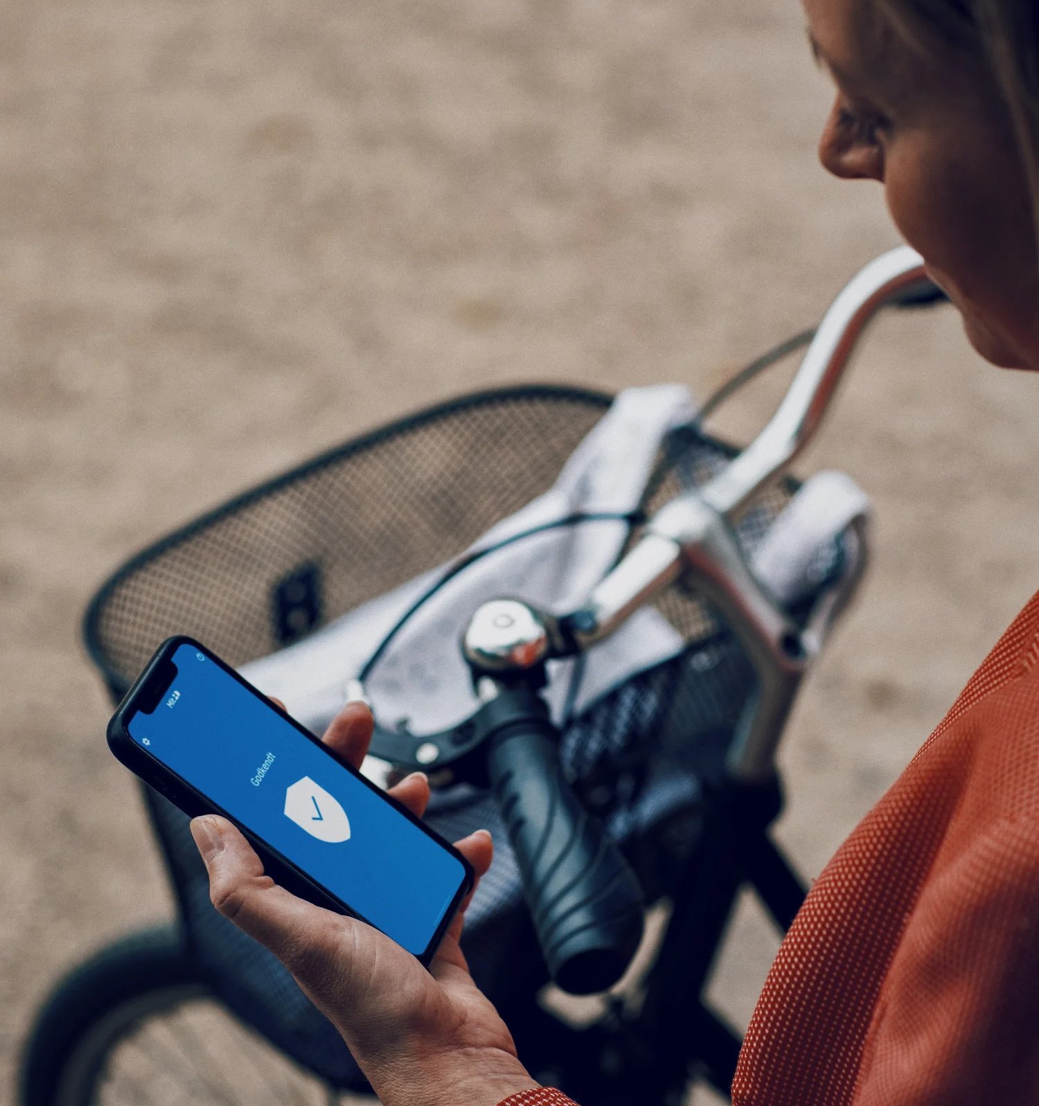 En person håller i en smartphone med en blå skärm som visar en sköld-ikon, bredvid en cykel med en flätad korg.