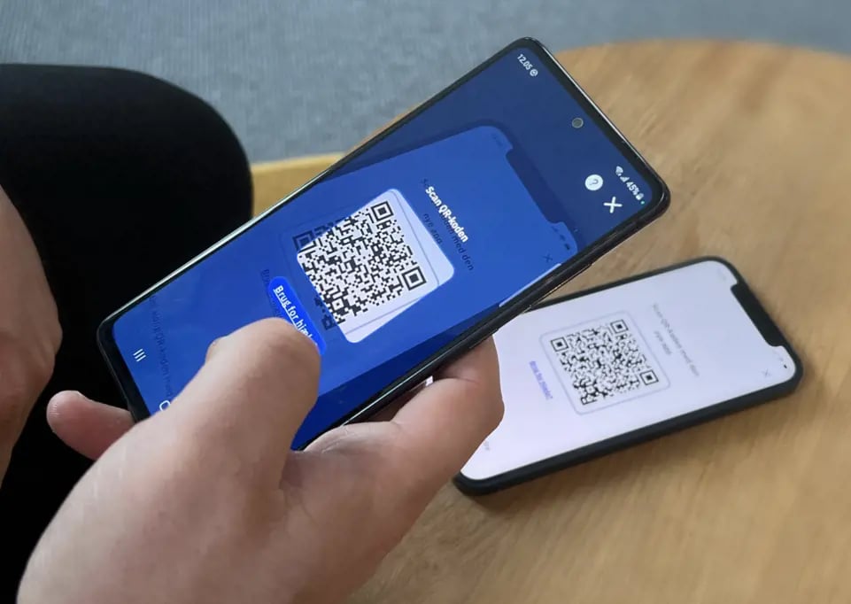 En person håller i en smartphone som visar en QR-kod, medan en annan telefon i bakgrunden visar en annan QR-kod på sin skärm.