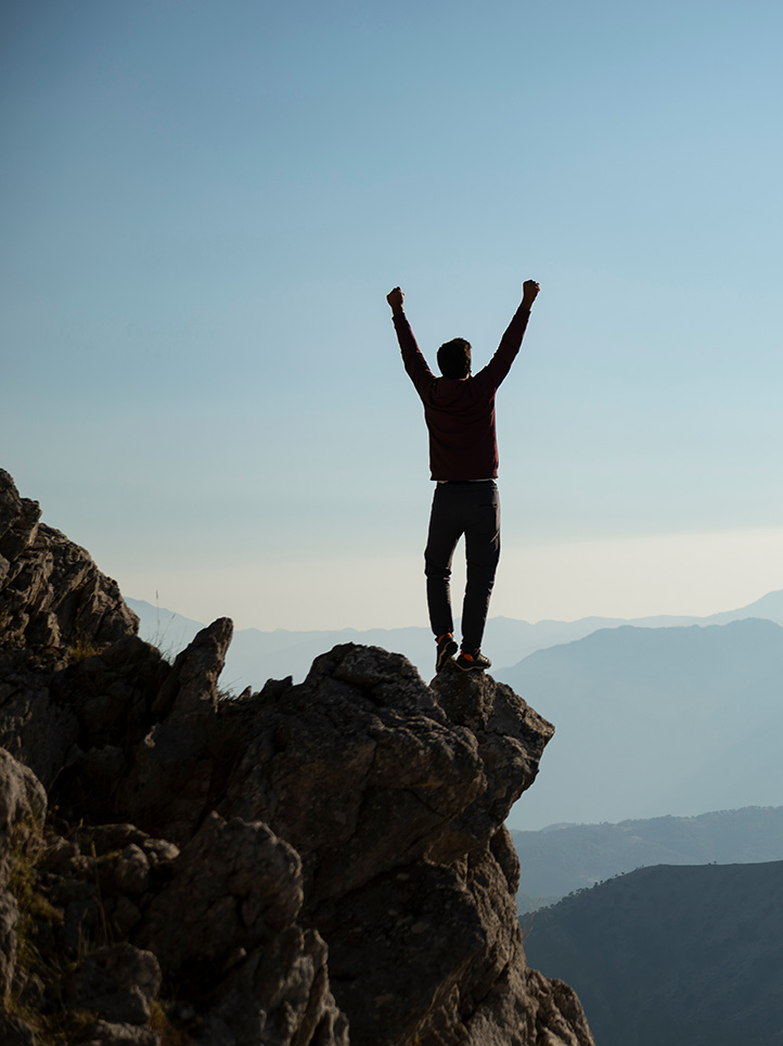 Man standing tall over cliff face with arms in the air celebrating.